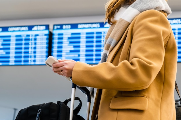 Woman traveler in stylish yellow coat with warm scarf holds mobile phone and suitcase standing again...