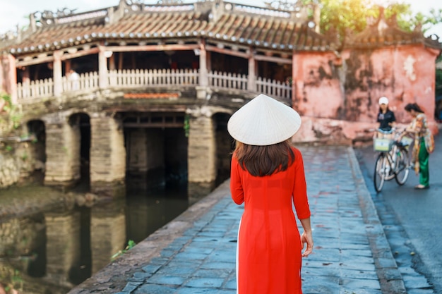 Woman traveler wearing Ao Dai Vietnamese dress sightseeing at Japanese covered bridge in Hoi An town Vietnam landmark and popular for tourist attractions Vietnam and Southeast Asia travel concept