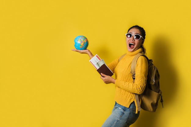 Woman traveler with suitcase, holding ball globe in the hand with passport and ticket on Yellow background. Portrait of smiling happy girl, concept of traveling around the world. Travel backpack