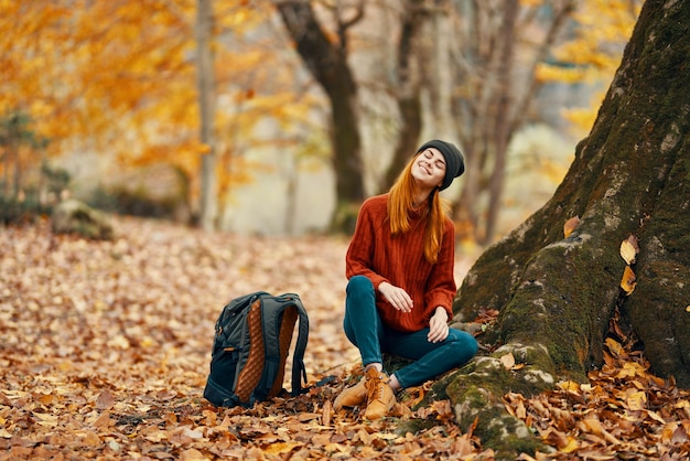 woman in trousers and boots with a backpack sits near a tree in the autumn forest fallen leaves High quality photo