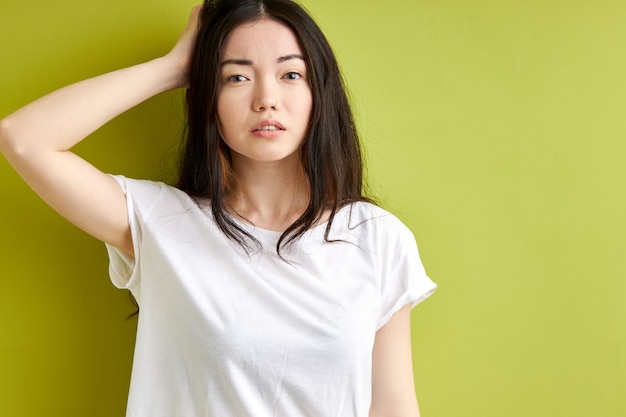 Woman try to remember something, stand thinking in contemplation touching hair and looking at camera, female in casual t-shirt isolated over green studio background