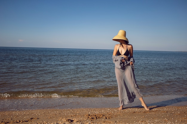 Woman in a tunic , a black swimsuit and a straw hat stands on the beach by the sea in summer on vacation