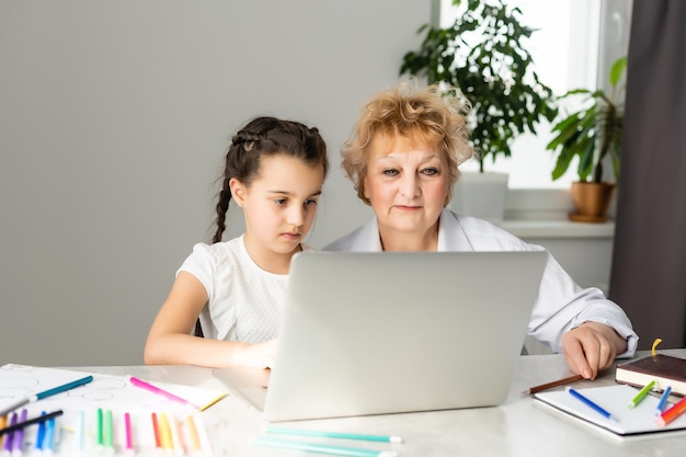 Woman tutor or foster parent mum helping cute caucasian school child girl doing homework sitting at table. Diverse nanny and kid learning writing in notebook studying at home.