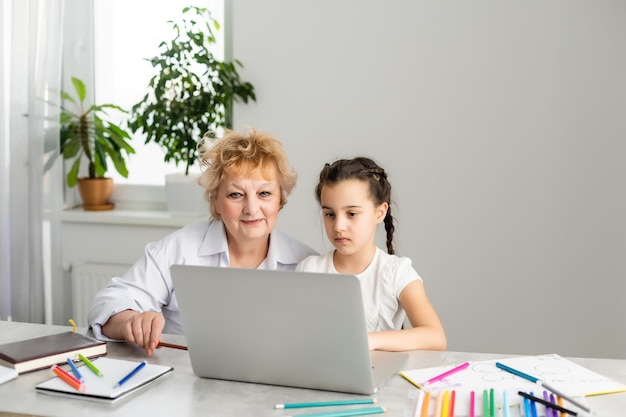 Woman tutor or foster parent mum helping cute caucasian school child girl doing homework sitting at table. Diverse nanny and kid learning writing in notebook studying at home.