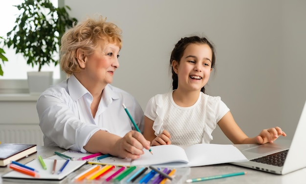 Woman tutor or foster parent mum helping cute caucasian school child girl doing homework sitting at table. Diverse nanny and kid learning writing in notebook studying at home.