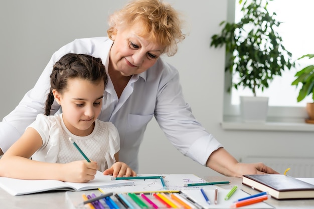 Woman tutor or foster parent mum helping cute caucasian school child girl doing homework sitting at table. Diverse nanny and kid learning writing in notebook studying at home.