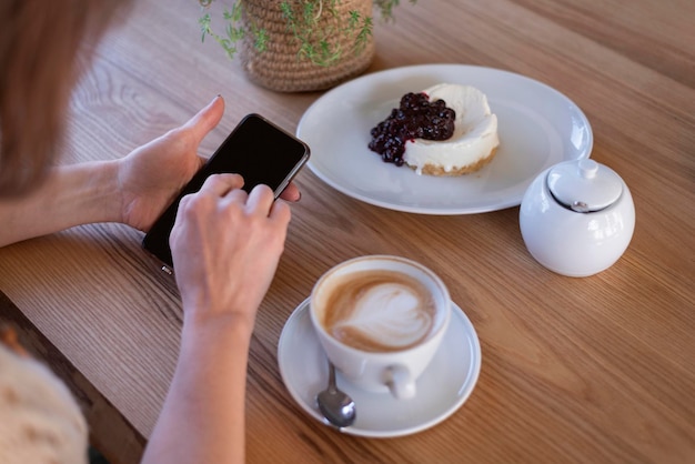 Woman uses mobile phone in cafe against table cappuccino coffee and dessert Smartphone in female hands