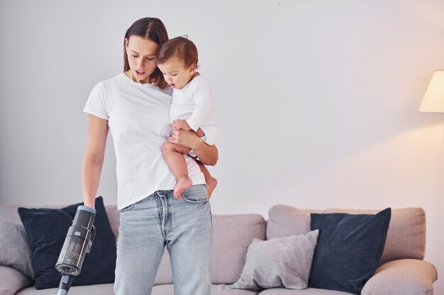 Woman uses vacuum cleaner Mother with her little daughter is indoors at home together