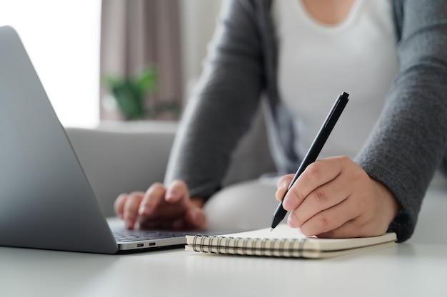 Woman using laptop and writing on notepad with a pen working at home office or workplacexD