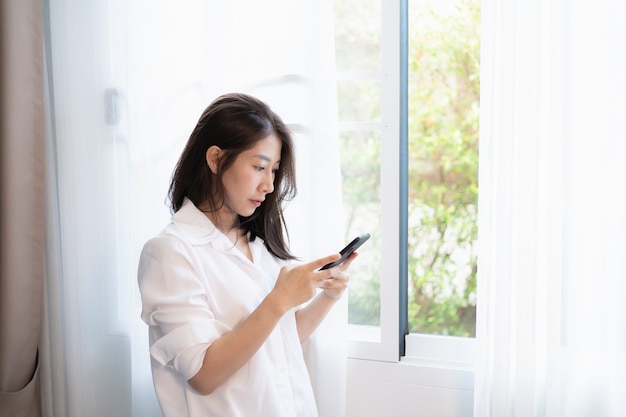 Woman using mobile phone and standing at the window in the bedroom