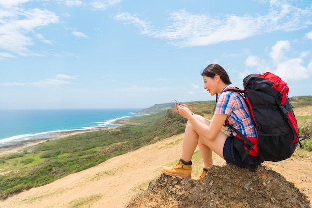 woman using mobile phone with family and friends chatting contact when hiking carrying backpack sitting on the hill top view coast landscape.