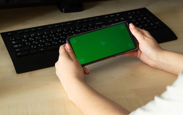 Woman using mobile smart phone with green screen on table with computer and keyboard