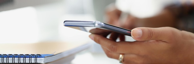 Woman using modern smartphone while sitting at table with spiral notebook in office