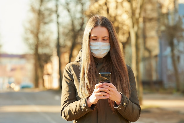 A woman using a smartphone wears a medical face mask to avoid the spread coronavirus on a city street. A girl with a surgical mask on the face against COVID-19.