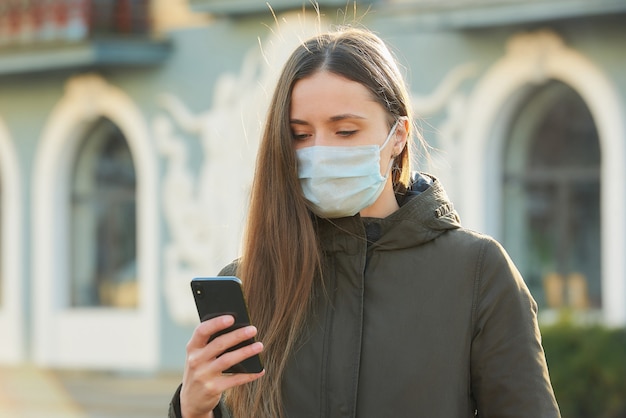 A woman using a smartphone wears a medical face mask to avoid the spread coronavirus on a street. A girl with a surgical mask on the face against COVID-19.