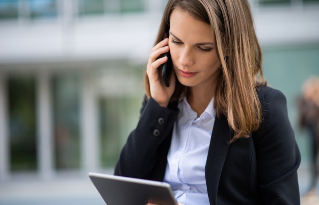 Woman using a tablet while talking on her mobile cell phone in front of her office