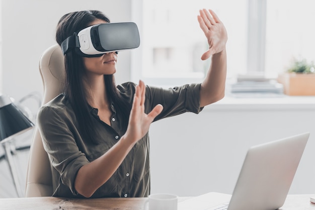 Woman in VR glasses. Confident young woman in virtual reality headset pointing in the air while sitting at her working place in office