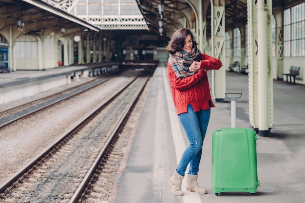 Woman waits train at railway station, dressed in knitted sweater and jeans, stands near green bag, looks at watch.