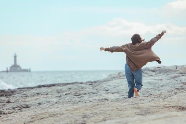 Woman walking by rocky sea beach in wet jeans lighthouse on background