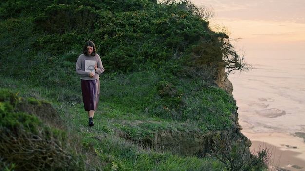 Woman walking coast hill at gloomy evening weather holding album with drawings