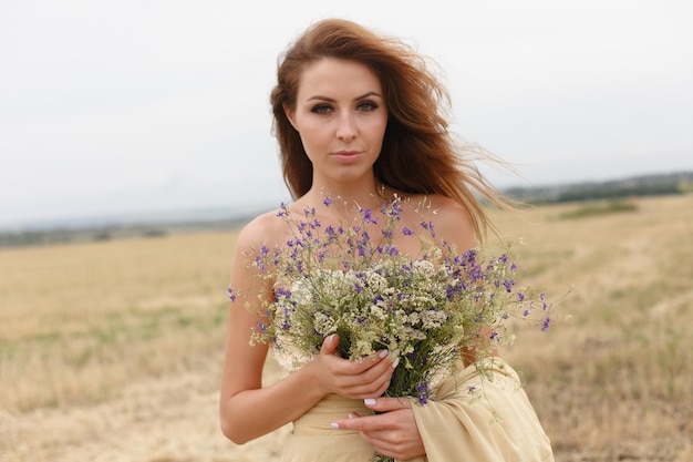 Woman walking in golden dried grass field Natural portrait beauty Beautiful girl in a wheat field Young woman in a beige dress holding a bouquet of wildflowers