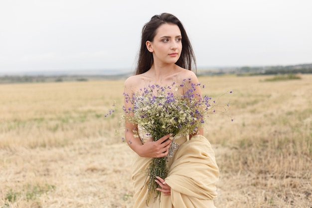 Woman walking in golden dried grass field Natural portrait beauty Beautiful girl in a wheat field Young woman in a beige dress holding a bouquet of wildflowers