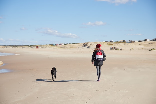 Woman walking with dog on the beach in winter under cloudy sky.