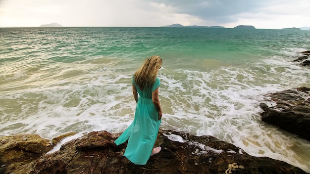 Woman walks on rock of sea reef stone stormy cloudy ocean Blue swimsuit dress tunic Concept rest tropical resort coastline tourism summer holidays