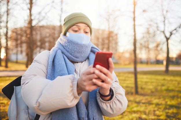 Woman in warm clothing and in mask using navigator on her mobile phone while walking in the city