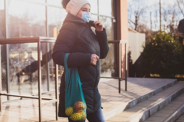 Woman in warm coat and knitted hat after shopping holding handmade mesh bag with fruits Sunny winter morning in front of supermarket