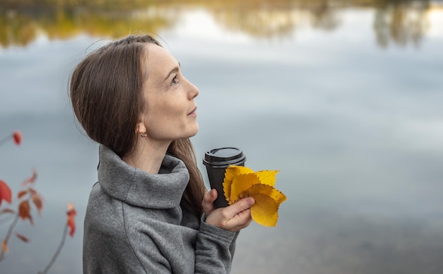 Woman in warm gray dress is walking through the autumn forest along the river with a cup of coffee Nature walks to think in silence and calm