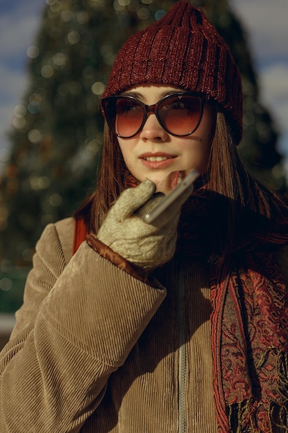 Woman in warm outerwear and sunglasses recording voice audio message on smartphone