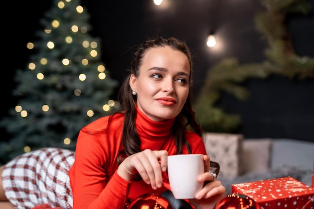 Woman in warm stylish clothes poses on bed in decorated room. Beauty in red sweater drinks coffee. Fest spirit.