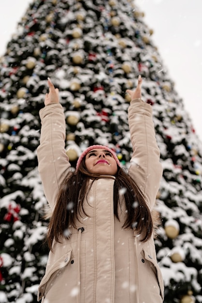 Woman in warm winter clothes standing by the big christmas tree outdoors, arms up, snow falling