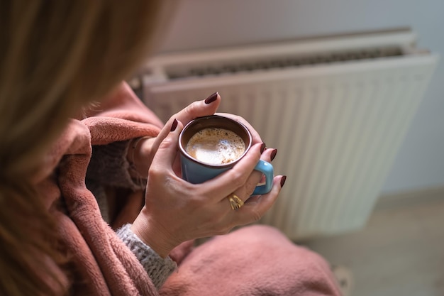 Woman warming up with a cup of coffee next to a radiator because of the cold of winter and energy savings