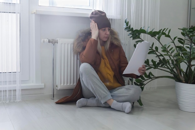 A woman warmly dressed in a brown jacket and hat is sitting near the heater with bills in her hands The concept of a crisis and a large set for utilities