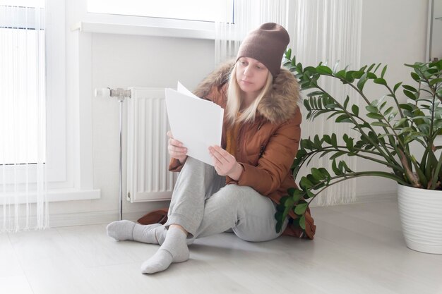 A woman warmly dressed in a brown jacket and hat is sitting near the heater with bills in her hands The concept of a crisis and a large set for utilities