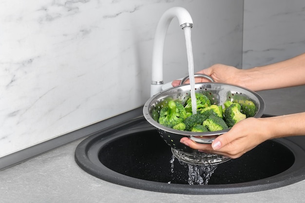 Woman washing fresh green broccoli in metal colander under tap water closeup view Space for text