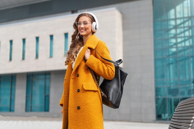 Woman Wear Eyeglasses Listen Music Headphone Walking Outdoor City Street Dressed Stylish Yellow Coat And Black Backpack Smile Caucasian Female 30s Enjoy Podcast Or Audio Books Outside