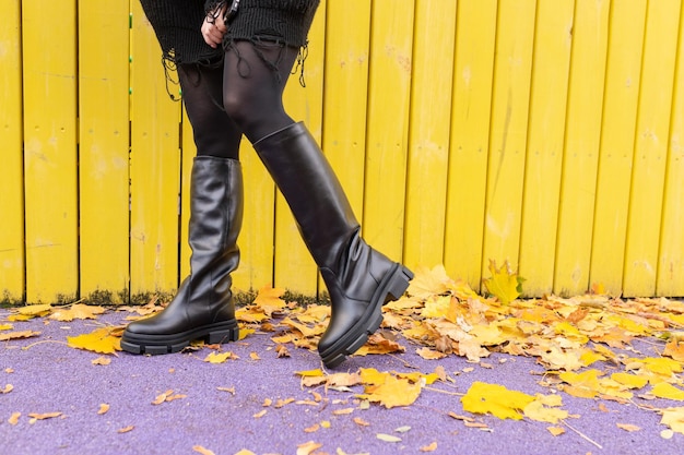 A woman wearing black boots stands in front of a yellow wooden fence.