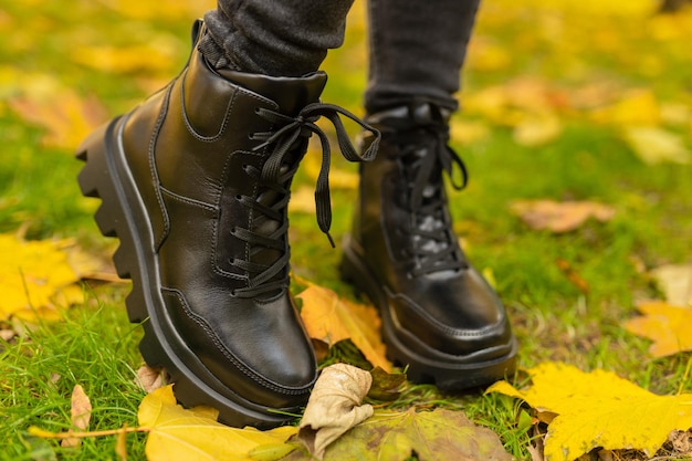 A woman wearing black boots stands in the grass with yellow leaves on the ground.