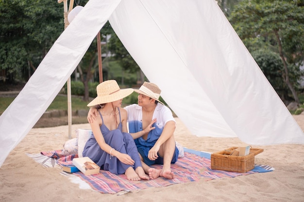 Woman wearing blue dress and straw hat and her boyfriend sitting under the white tent on a beach