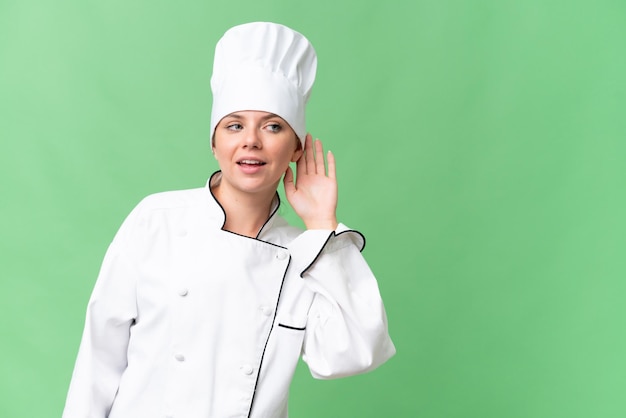 A woman wearing a chef hat holds her hand to her ear and talks to the camera.