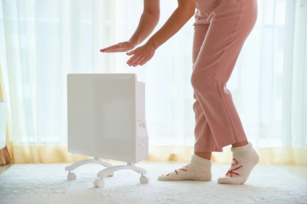 Woman wearing cozy comfort soft knitted socks get warming hands over modern white portable electric heater at home