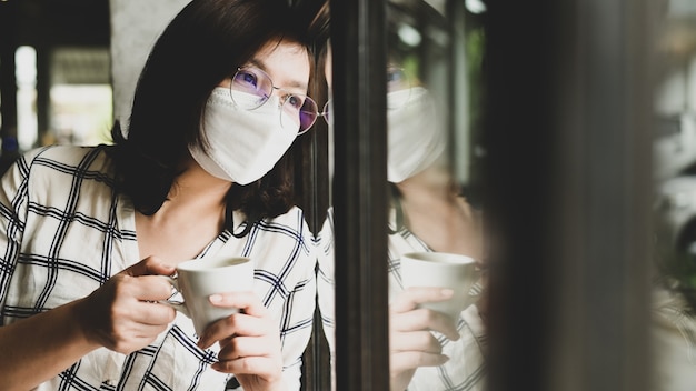 A woman wearing a medical mask with a coffee cup is looking out the window, she is tired of quarantine during the coronavirus, working from home, quarantine, new normol.