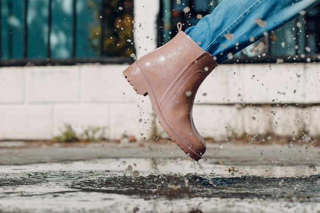 Woman wearing rain rubber boots walking running and jumping into puddle with water splash and drops in autumn rain