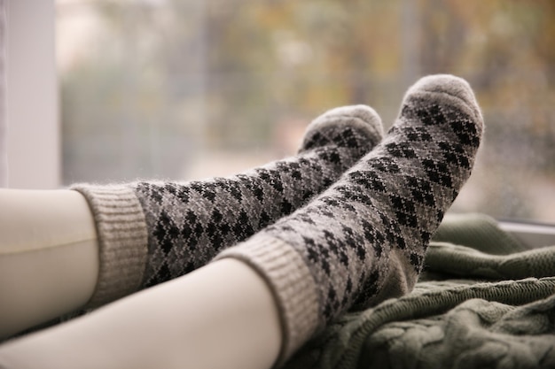 Woman wearing warm socks on knitted plaid near window closeup Cozy season