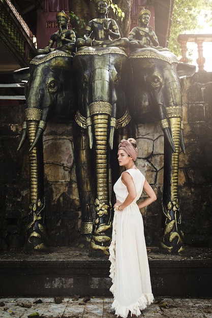 Woman wearing white robe dress and headscarf posing beside three headed elephant statue