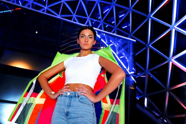 A woman wearing a white top and blue jeans stands in front of a colorful flag.