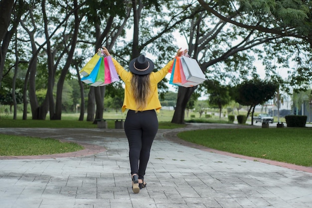 Woman wearing yellow jacket walking in a garden with multicolored shopping bags Shopping concept